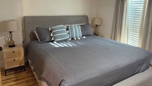 Una cama bien hecha con colcha gris, dos mesitas de noche con lámparas y una ventana con cortinas.