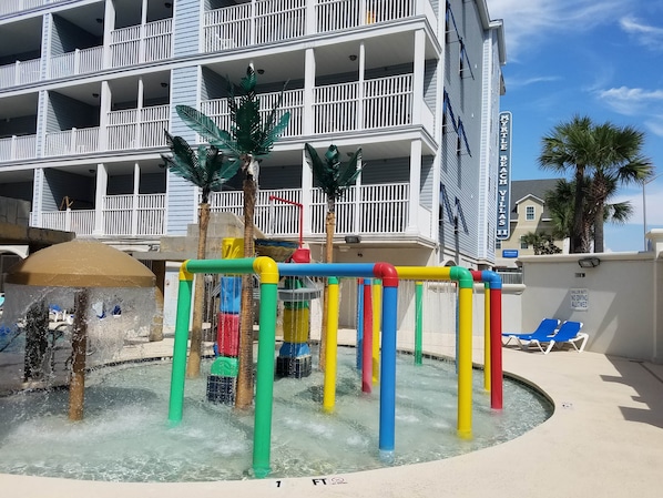 A water play area with colorful structures, a pool, and a building with balconies in the background.