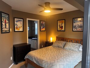 Une chambre à coucher avec un lit, une table de chevet, une commode et un ventilateur de plafond. Des tableaux encadrés ornent les murs.