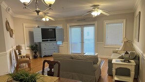 A living room with a sofa, a TV, a dining table with chairs, a lamp, and a ceiling fan. - Luxury 3BR 2.5 B Condo Just Steps from the Ocean! (Tybee Island)