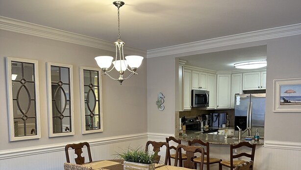 A dining area with a wooden table and chairs, a chandelier, and a kitchen in the background. - Luxury 3BR 2.5 B Condo Just Steps from the Ocean! (Tybee Island)