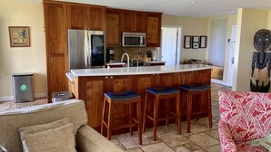 Une cuisine avec des meubles de rangement en bois, un réfrigérateur en acier inoxydable, un four à micro-ondes et un évier. On y trouve trois tabourets de bar et une chaise rouge à motifs.