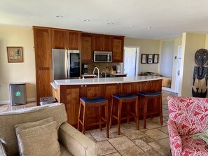 A kitchen with wooden cabinets, a stainless steel refrigerator, a microwave, and a sink. There are three bar stools and a red patterned chair. - Wailea Ekahi 54A: Remodeled & Panoramic Ocean View. (Kihei)