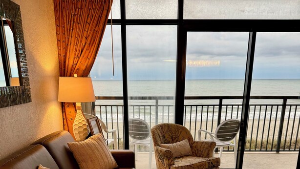 A cozy living room with a leather sofa, patterned armchair, and a round coffee table. A large window offers a view of the beach and ocean. - Gorgeous direct oceanfront- end unit in highly desirable location! 
 (North Myrtle Beach)