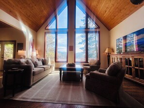 A cozy living room with a high ceiling, large windows, a fireplace, and a television.