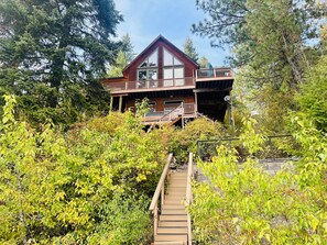 A two-story wooden cabin with a balcony, surrounded by dense foliage and a wooden staircase leading to the entrance. - Family-friendly Lakefront Home w/Kayaks, Paddleboards & More-Close to Silverwood (Hayden)