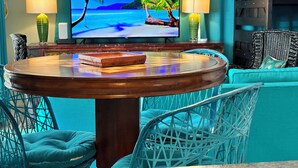 Ein Zimmer mit einem runden Holztisch, zwei Stühlen, einer Couch, einem Fernseher, der eine tropische Strandlandschaft zeigt, und zwei Lampen.