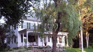 A white colonial-style house with a front porch, a stone retaining wall, and a large tree in the yard. - Renovated 1854 Sea Captain's House w/Harbor Views in Deer Isle Village (Deer isle)