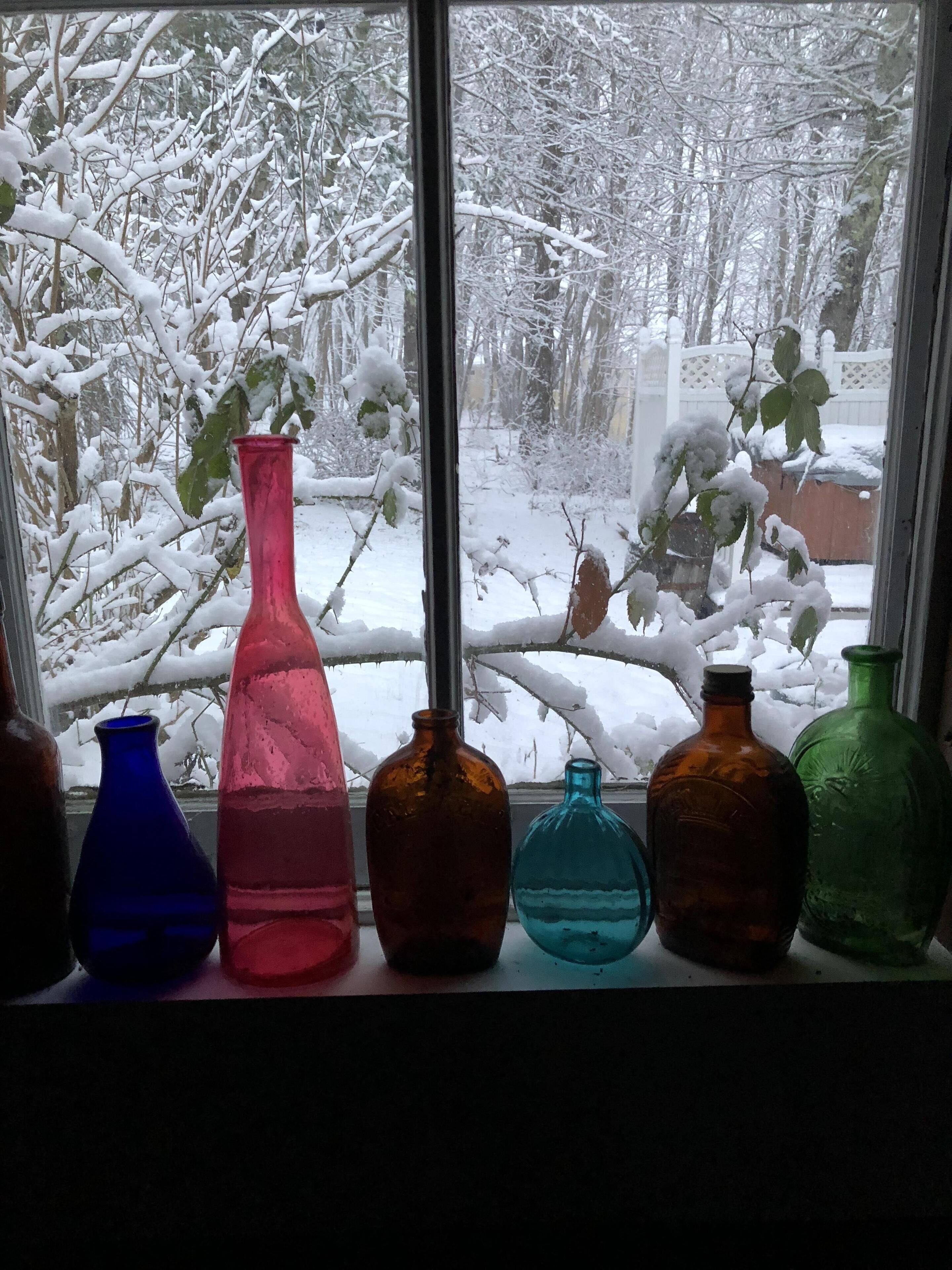 A window with six colored glass bottles on a sill, overlooking a snowy landscape.