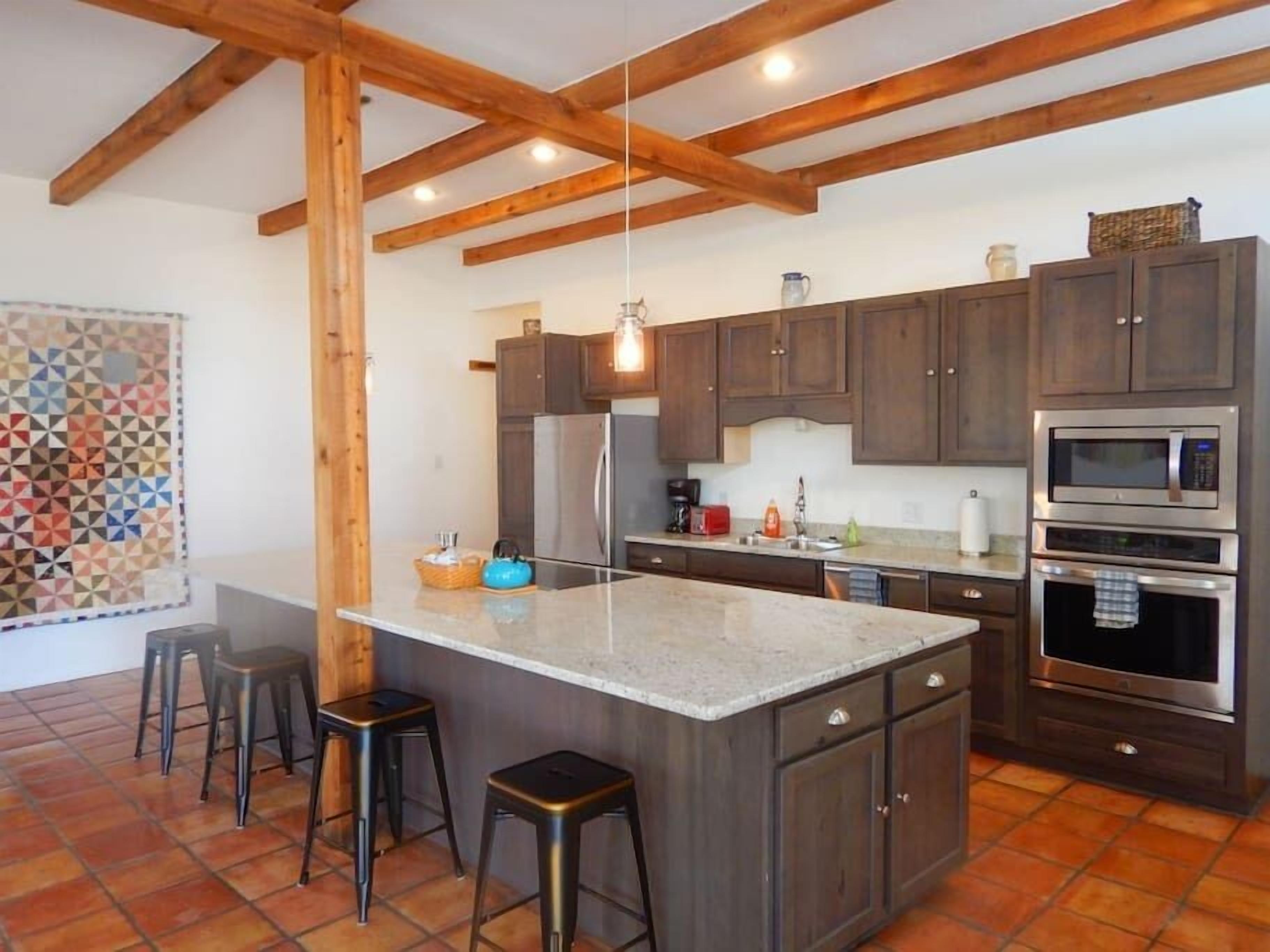 Une cuisine avec des poutres en bois, des meubles de couleur sombre, un îlot central avec des tabourets et des appareils électroménagers modernes.