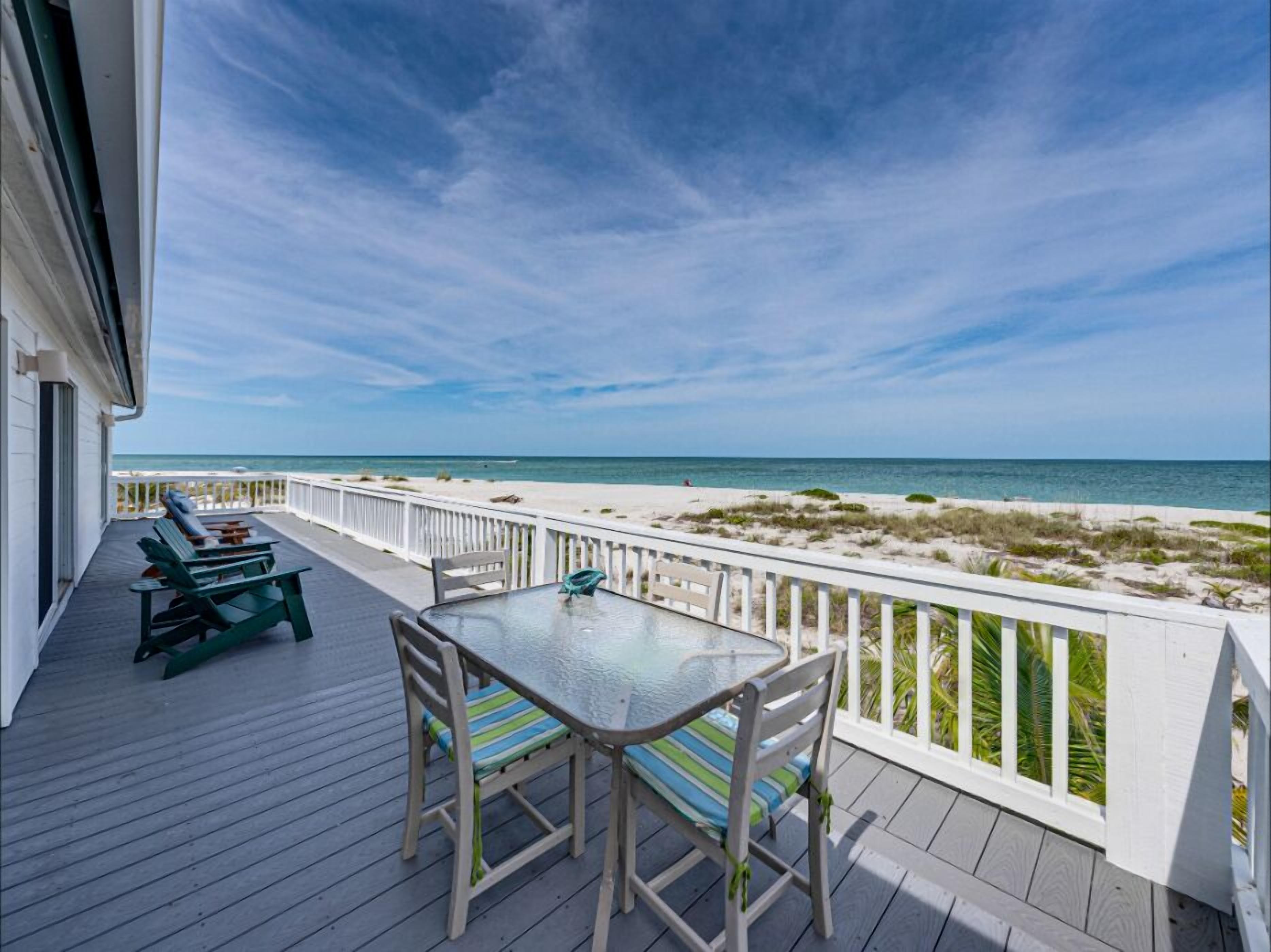 Une terrasse en bois avec une table et des chaises en verre, donnant sur une plage de sable blanc et un ciel bleu azur.
