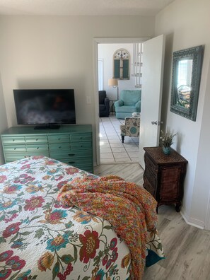 A bedroom with a bed, a dresser, a television, a door leading to another room, and a mirror on the wall.