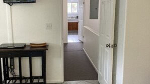 A hallway with a tiled floor, a table with a basket, a mounted TV, and a door leading to a kitchen area. - 1 Mile from Pipeline Surf Break! (Haleiwa)