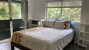 A bedroom with a bed, a window with blinds, a chair, and a small table. - 1 Mile from Pipeline Surf Break! (Haleiwa)