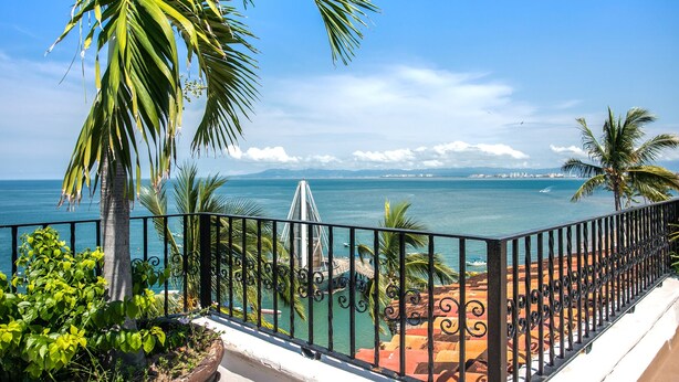 A balcony with a view of the sea, palm trees, and a distant cityscape.