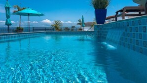 A clear blue swimming pool with a tiled edge, surrounded by umbrellas and potted plants.