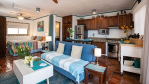 A cozy living room with a blue sofa, a coffee table with flowers, and a kitchen with wooden cabinets and a refrigerator.