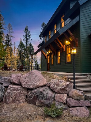 A two-story building with a stone foundation, wooden beams, and multiple windows with warm lighting. - Spectacular Views of Swan Mountain Range~Spacious Home~Perfect for Entertaining (Seeley Lake)
