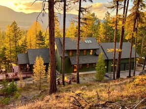 A cabin with a deck, surrounded by tall trees and a mountain backdrop.