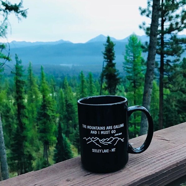 A black mug with a quote on a wooden deck overlooking a forest and mountains. - Swan Mountain Views~Private 27-Acre Retreat Near Seeley Lake (Seeley Lake)