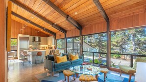 A modern interior with a wooden ceiling, a kitchen area, a seating area with a glass table, and large windows offering a view of the outdoors. - Pebble Beach perfection. Peaceful, Ocean view property to rest, golf, reset. (Del Monte Forest)