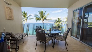 Ein Balkon mit rundem Tisch und vier Stühlen, mit Blick auf einen Strand mit Palmen und dem Meer.