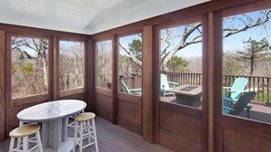 Un espace extérieur abrité, doté d’une terrasse en bois, d’une table ronde et de deux tabourets, et offrant une vue sur les arbres et le ciel environnants.