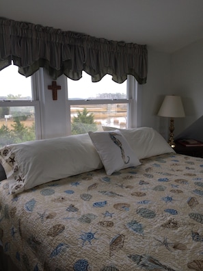 A bedroom with a bed, a window with a view of a landscape, a cross on the wall, and a lamp on a bedside table.