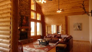 A log cabin interior with a stone fireplace, leather sofa, and a large window. - Premium Mountain Retreat On Over 2 Acres With Sweeping Views-hot Tub/Pool Table (Ruidoso)