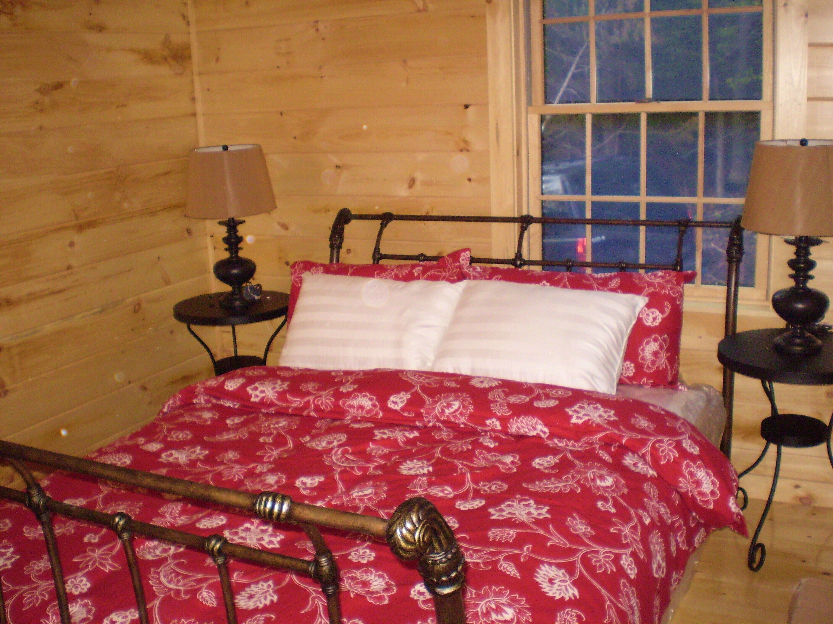 Un dormitori de cabana de fusta amb un llit de metall, dues taules de nit amb làmpades i una finestra amb vista als arbres.