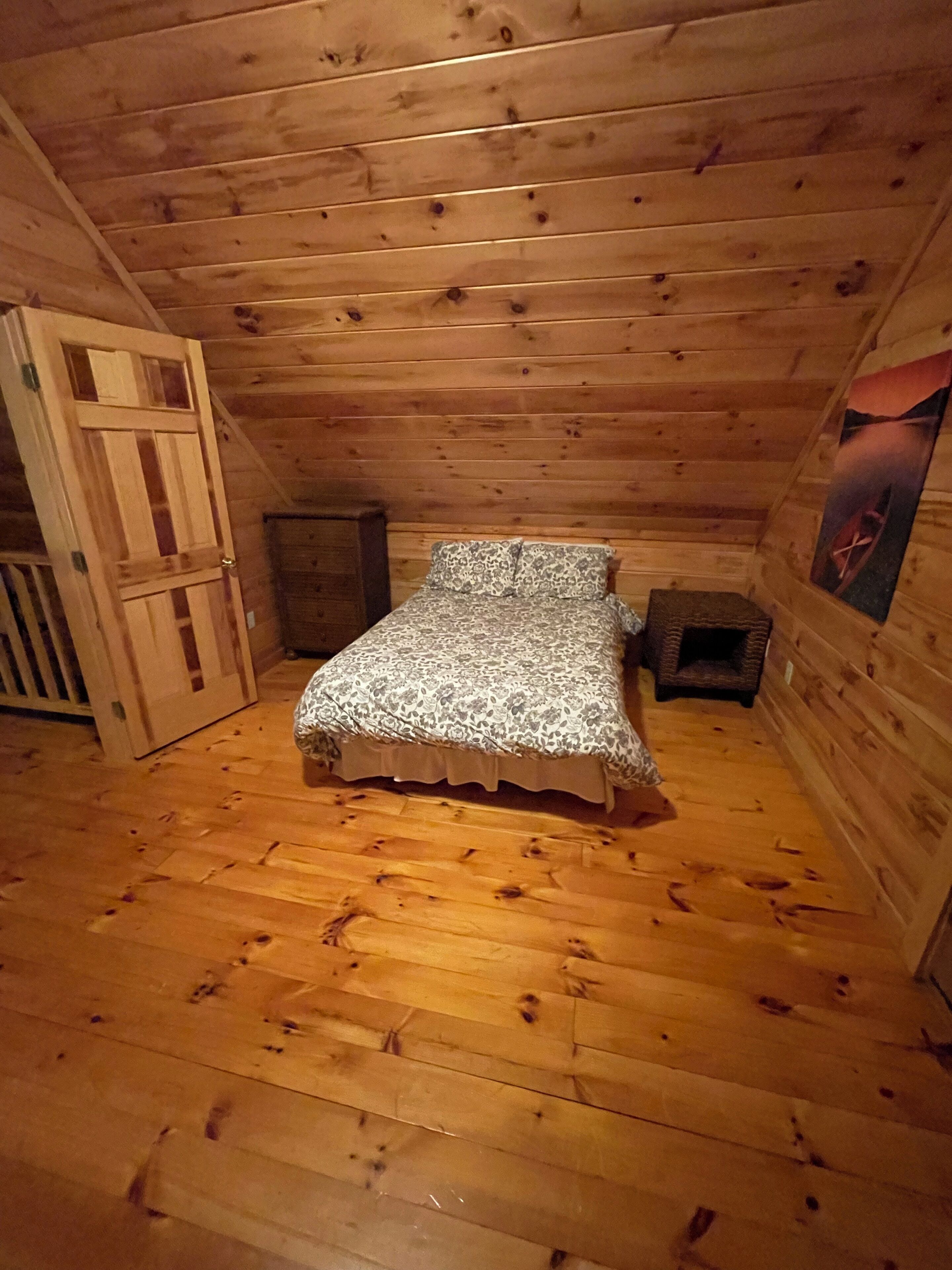 A small, cozy room with wooden walls and a sloped ceiling. It features a single bed with patterned bedding, a small wooden cabinet, and a door leading to another room.
