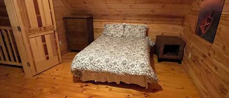 A small, cozy room with wooden walls and a sloped ceiling. It features a single bed with patterned bedding, a small wooden cabinet, and a door leading to another room.