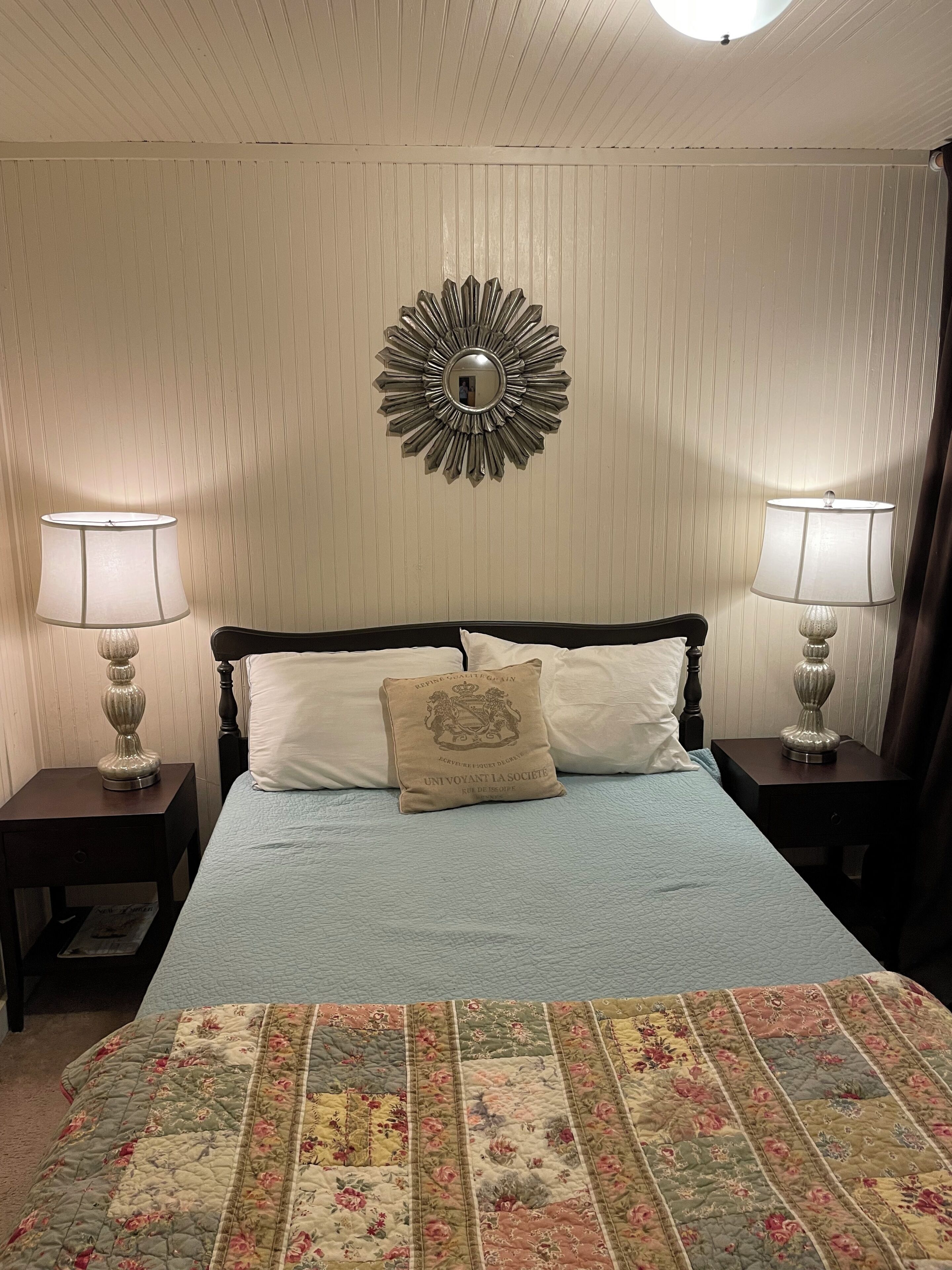 A neatly made bed with a floral quilt, two bedside lamps, a decorative mirror, and a pillow with a crest.