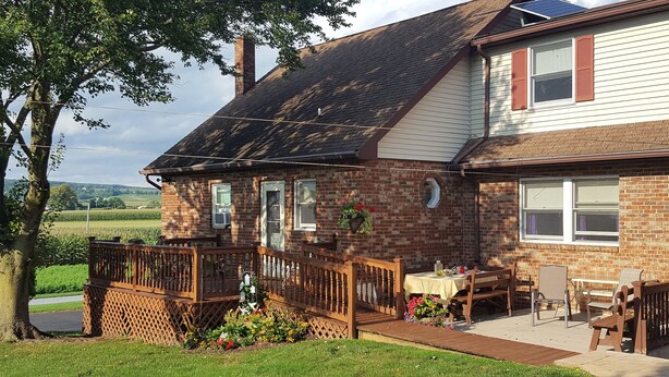 A two-story brick house with a wooden deck, a small patio with a table and chairs, and a garden with flowers. - Scenic View Lodging ~ A Country Suite with Country Charm. (Gap)