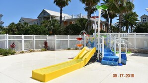 Un espace de jeux aquatiques avec des toboggans et des jets d’eau, entouré d’une clôture blanche et de palmiers.