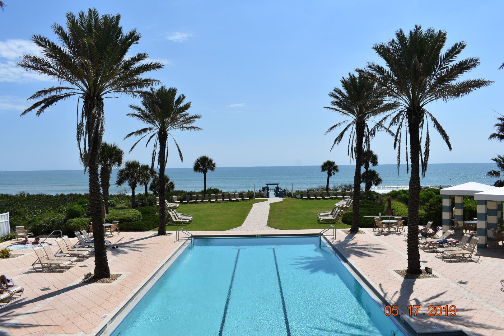 A pool area with lounge chairs and palm trees, overlooking a beach and ocean.