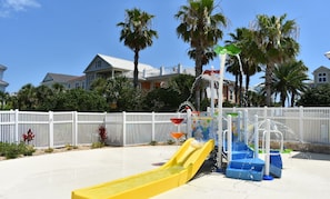 A water play area with slides and water jets, surrounded by a white fence and palm trees. - 5 Bedroom Luxury Gated Ocean View Home with Amenities (Palm Coast)