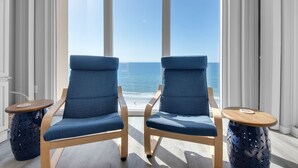Zwei blaue, gepolsterte Sessel mit Holzrahmen und runde Holztische mit gelochten Seiten stehen vor einem großen Fenster mit Blick auf den Ozean.