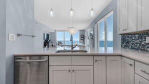 Eine moderne Küche mit Geschirrspüler aus Edelstahl, weißen Schränken und einer großen Kücheninsel mit Spüle. Drei Pendelleuchten beleuchten die Insel, und ein großes Fenster bietet Blick aufs Meer.