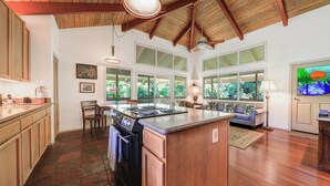 A spacious kitchen with a central island, wooden cabinets, and a large window overlooking greenery. - Paddle to the Beach from a Secluded Riverfront Eden-A/C-Central to Hanalei&Ke'e (Hanalei)