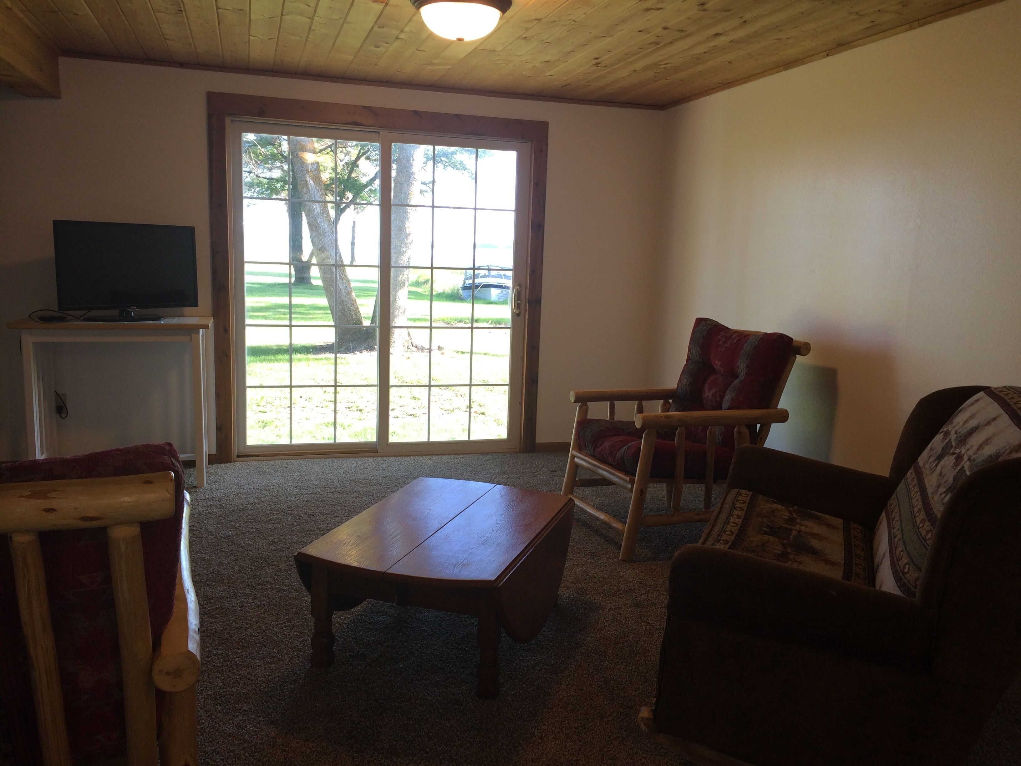 A living room with a wooden table, a red cushioned chair, a sofa, a television on a stand, and a large sliding glass door leading to a grassy area.