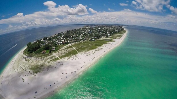 Vista aérea de una isla costera con una playa larga y arenosa, aguas turquesas cristalinas y una zona residencial densa con casas y vegetación.