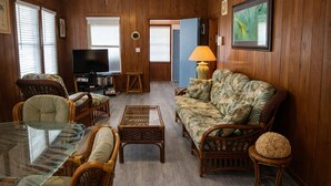 Una sala de estar con televisor, muebles de mimbre, una mesa de centro de vidrio y un sofá estampado con motivos tropicales.