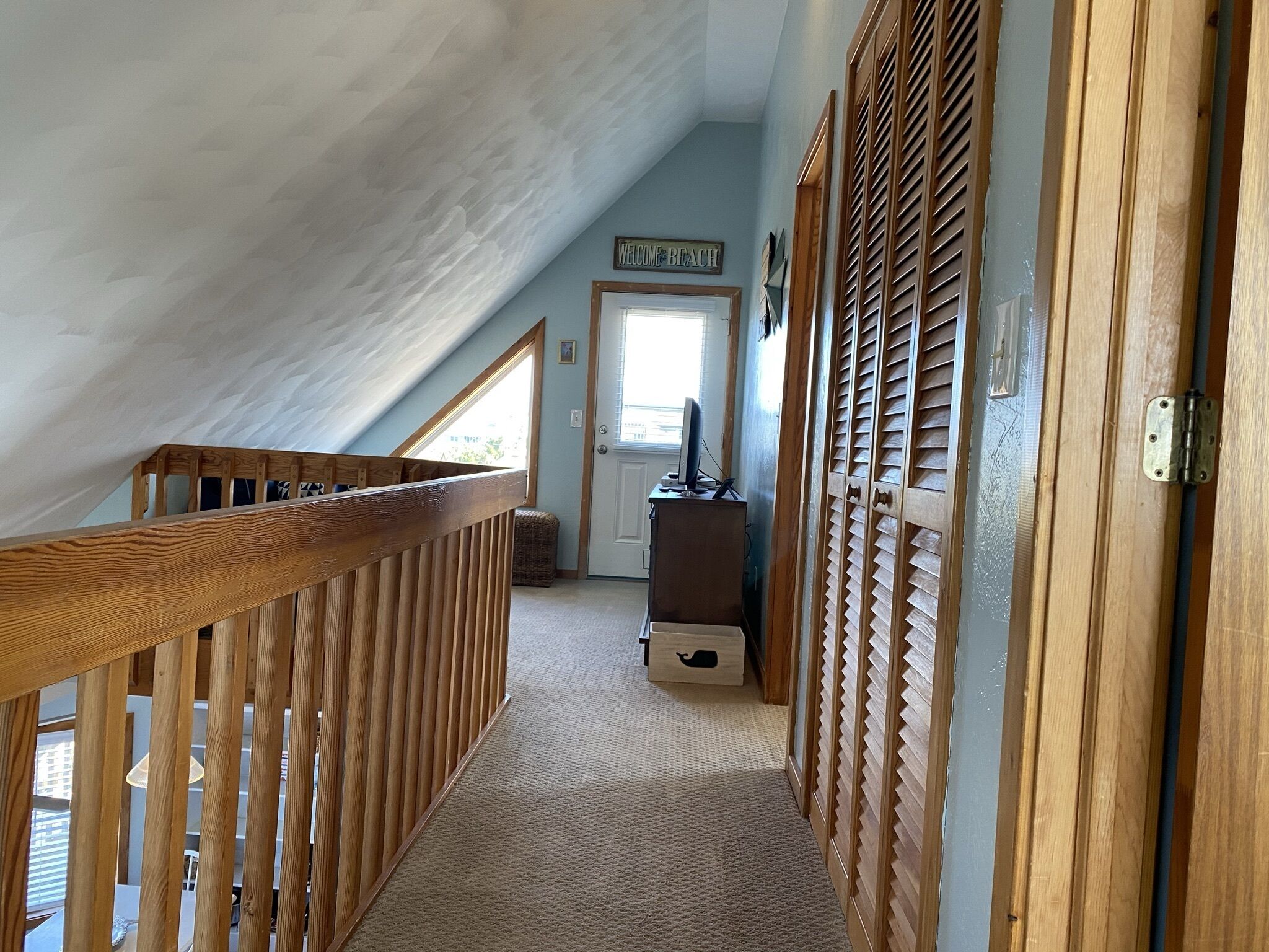A narrow hallway with a wooden railing, a door with a 'Welcome Beach' sign, and a small desk with a computer.