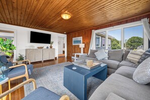 Ein modernes Wohnzimmer mit Holzboden, einem an der Wand montierten Flachbildfernseher, einer Sofagarnitur, einem Couchtisch und Blick ins Freie durch große Fenster.