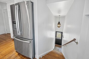 A modern kitchen with a stainless steel refrigerator, wooden flooring, and a staircase leading to a dark door.