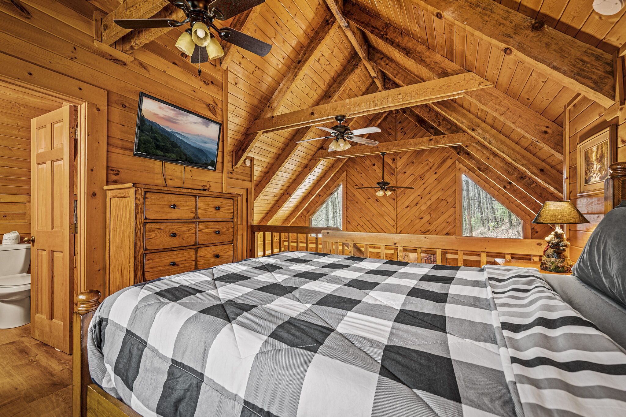 A cozy bedroom with a checkered bedspread, wooden ceiling beams, and a television on the wall.