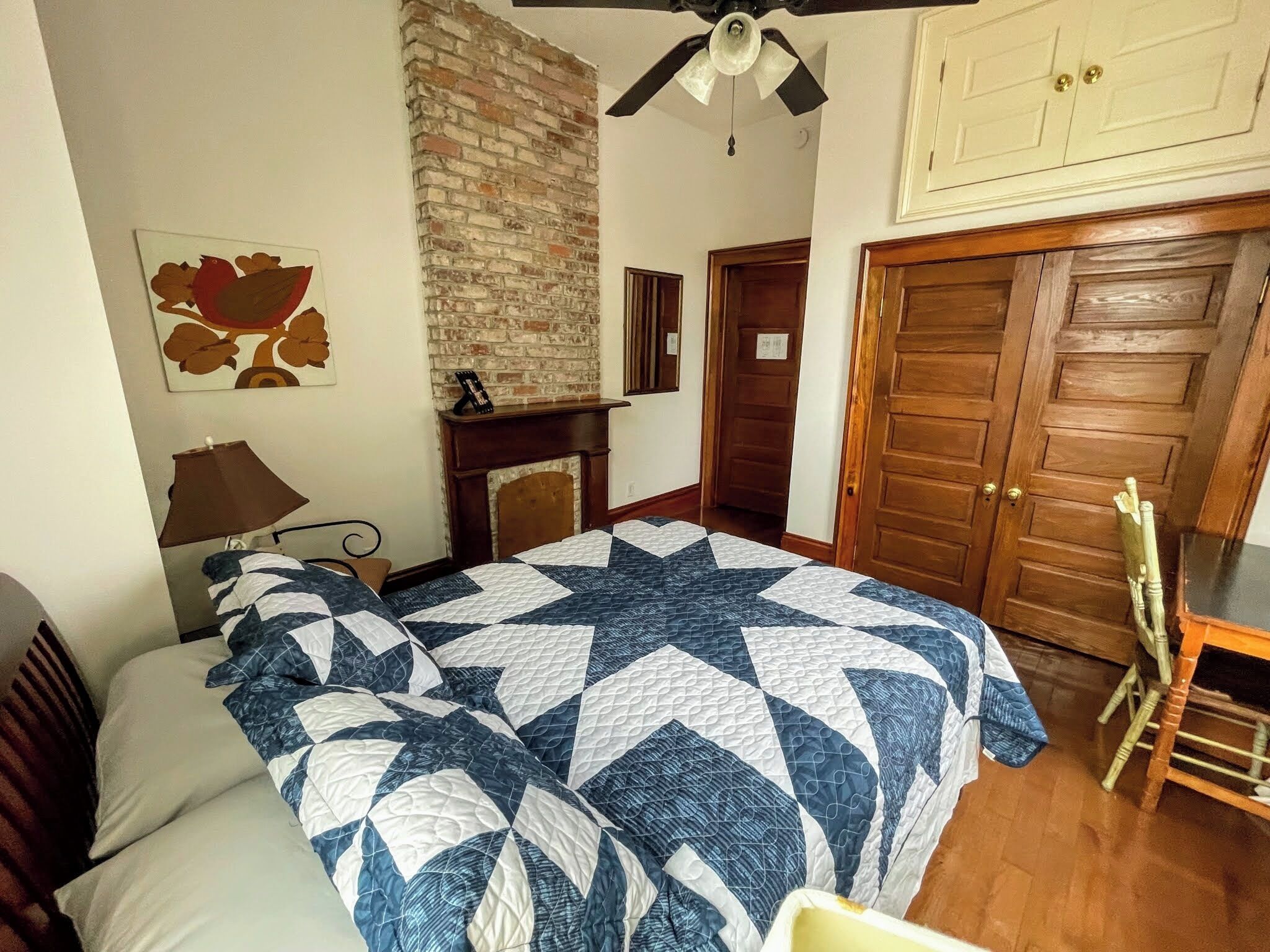 Een slaapkamer met een bed, nachtkastje, lamp, open haard, houten deur en plafondventilator.