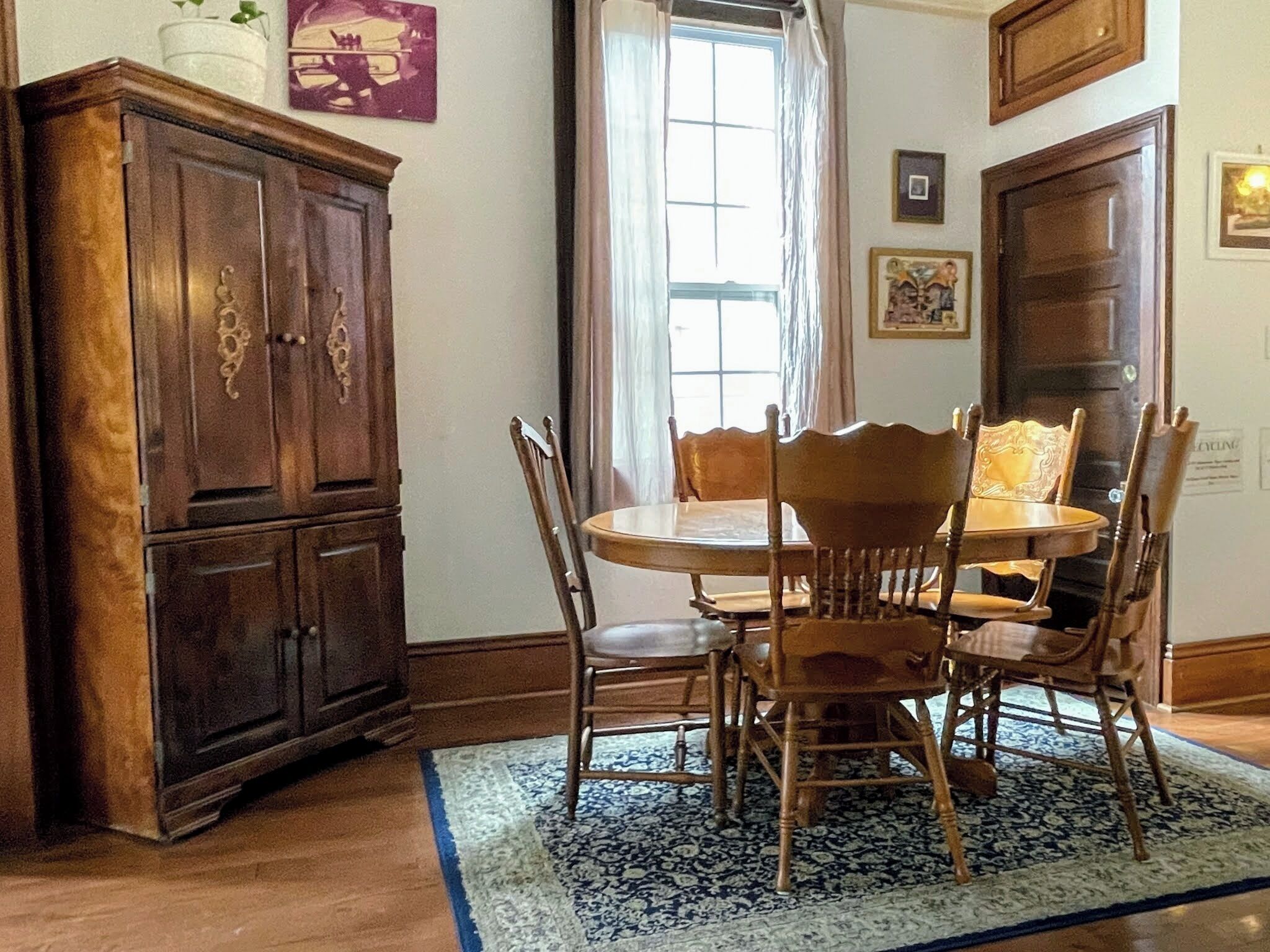 Een eethoek met een ronde tafel, zes houten stoelen, een donker houten kast en een tapijt met patroon.