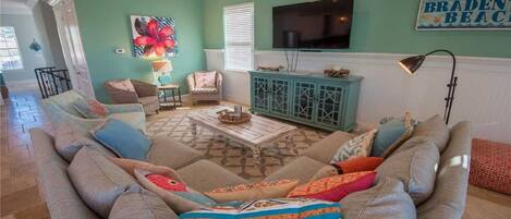 A living room with a teal wall, a large sofa with colorful cushions, a coffee table, and a TV mounted above a teal cabinet.